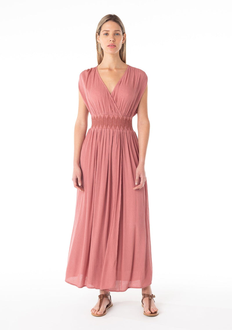 [Color: Terracotta] A front facing image of a blonde model wearing a bohemian resort maxi dress in a soft terracotta pink. A sleeveless vacation style with a flowy long skirt, a side slit, an open back with tie closure, a v neckline in the front and back, and a smocked elastic waist with embroidered stitch detail. 
