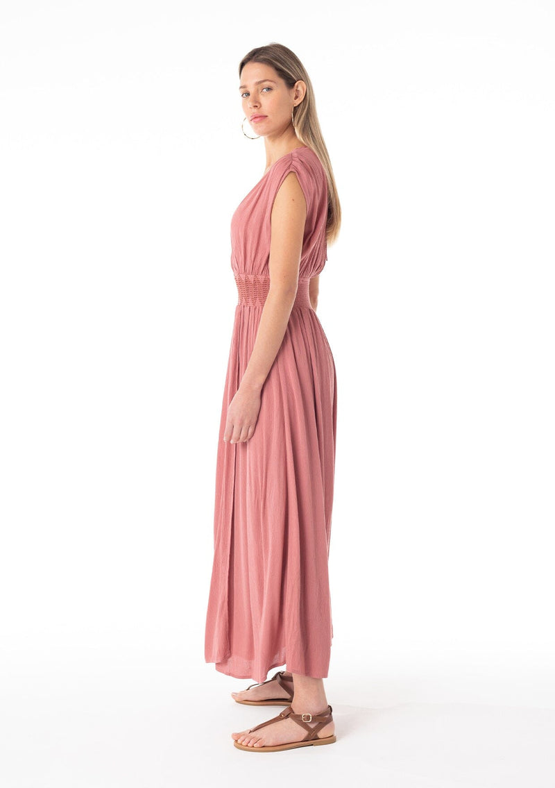 [Color: Terracotta] A side facing image of a blonde model wearing a bohemian resort maxi dress in a soft terracotta pink. A sleeveless vacation style with a flowy long skirt, a side slit, an open back with tie closure, a v neckline in the front and back, and a smocked elastic waist with embroidered stitch detail. 