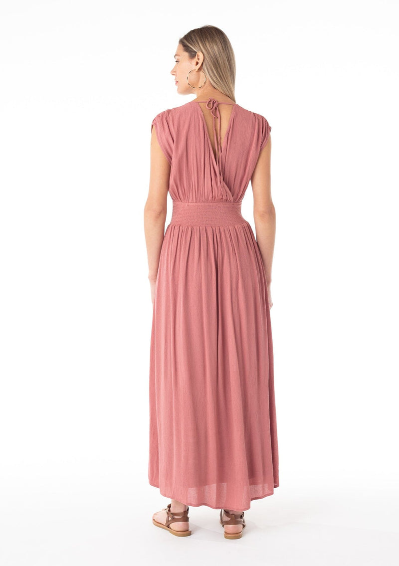 [Color: Terracotta] A back facing image of a blonde model wearing a bohemian resort maxi dress in a soft terracotta pink. A sleeveless vacation style with a flowy long skirt, a side slit, an open back with tie closure, a v neckline in the front and back, and a smocked elastic waist with embroidered stitch detail. 
