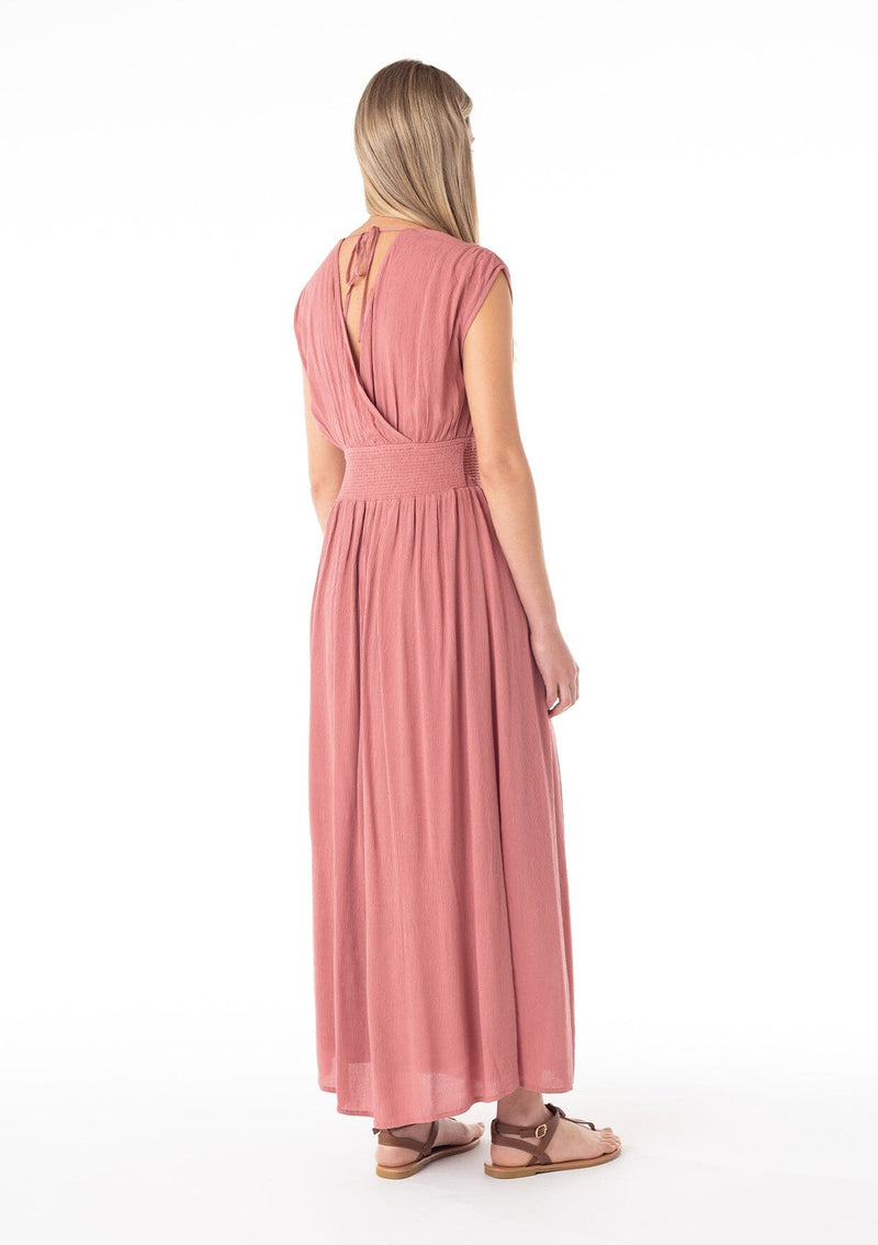 [Color: Terracotta] A side back facing image of a blonde model wearing a bohemian resort maxi dress in a soft terracotta pink. A sleeveless vacation style with a flowy long skirt, a side slit, an open back with tie closure, a v neckline in the front and back, and a smocked elastic waist with embroidered stitch detail. 