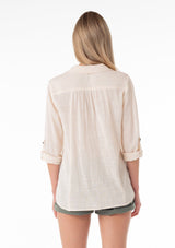 [Color: Natural] A back facing image of a blonde model wearing a natural, off white cotton shirt. With long rolled sleeved, a button tab sleeve closure, a button front, two front patch pockets, a tie front waist detail, and a high low hemline.