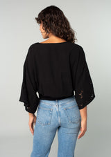 [Color: Black] A back facing image of a brunette model wearing a bohemian black embroidered eyelet top with billowy half length sleeves, a button front, and a v neckline. 