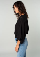 [Color: Black] A side facing image of a brunette model wearing a bohemian black embroidered eyelet top with billowy half length sleeves, a button front, and a v neckline. 