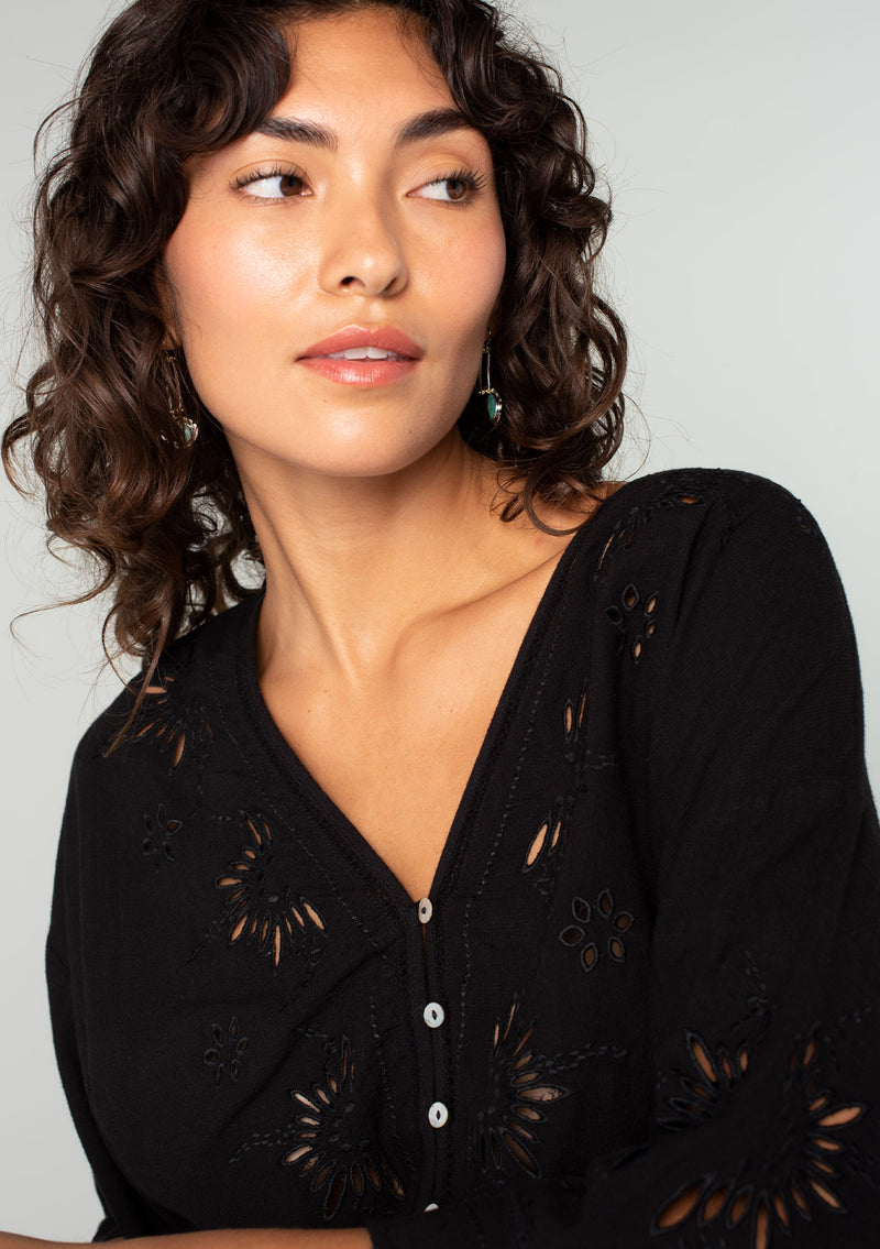 [Color: Black] A close up front facing image of a brunette model wearing a bohemian black embroidered eyelet top with billowy half length sleeves, a button front, and a v neckline. 