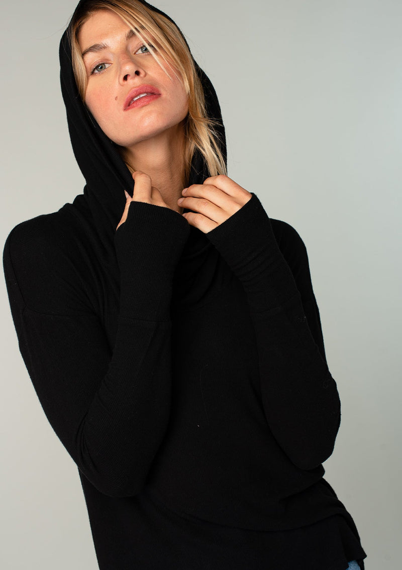 [Color: Black] A woman standing outside wearing a soft bamboo micro rib long sleeve top. Featuring an exaggerated cowl neckline that doubles as a hood, long sleeves with thumbhole accents, and breezy side vents.