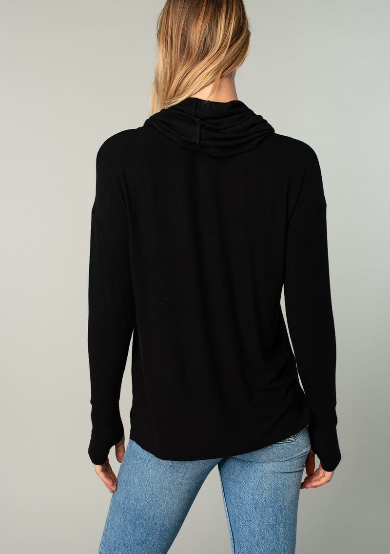 [Color: Black] A woman standing outside wearing a soft bamboo micro rib long sleeve top. Featuring an exaggerated cowl neckline that doubles as a hood, long sleeves with thumbhole accents, and breezy side vents.