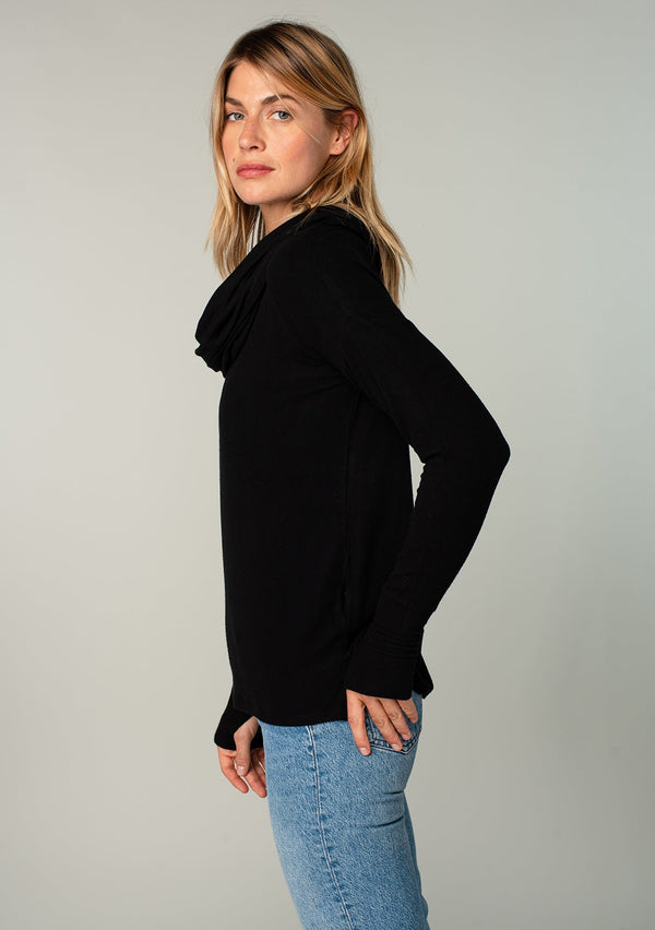 [Color: Black] A woman standing outside wearing a soft bamboo micro rib long sleeve top. Featuring an exaggerated cowl neckline that doubles as a hood, long sleeves with thumbhole accents, and breezy side vents.