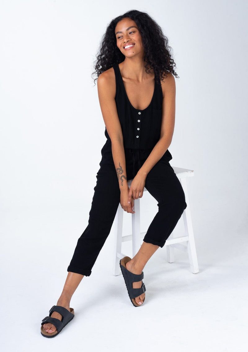 [Color: Black] A sitting front facing image of a brunette model wearing a classic bohemian one piece black jumpsuit. With a racer back, a button front top, side pockets, a cuffed wide leg, a scoop neckline, and an elastic waist with drawstring.