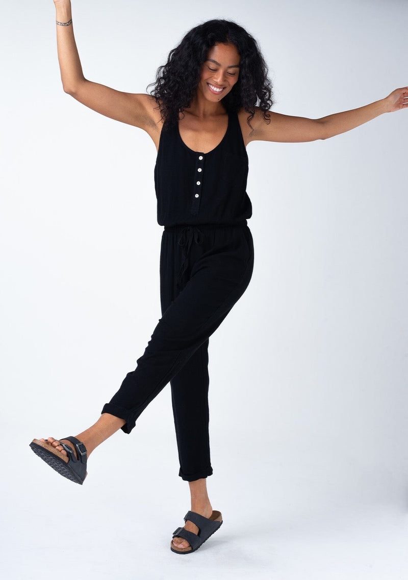 [Color: Black] A full body front facing image of a brunette model wearing a classic bohemian one piece black jumpsuit. With a racer back, a button front top, side pockets, a cuffed wide leg, a scoop neckline, and an elastic waist with drawstring.