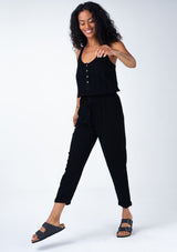 [Color: Black] A side facing image of a brunette model wearing a classic bohemian one piece black jumpsuit. With a racer back, a button front top, side pockets, a cuffed wide leg, a scoop neckline, and an elastic waist with drawstring.