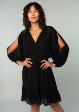 [Color: Black] A half body front facing image of a brunette model wearing a black bohemian mini dress in embroidered chiffon. With long split sleeves, a ruffle trimmed tiered skirt, a smocked elastic waist, a v neckline, and an open back with tassel tie closure. Perfect for weddings or date nights.