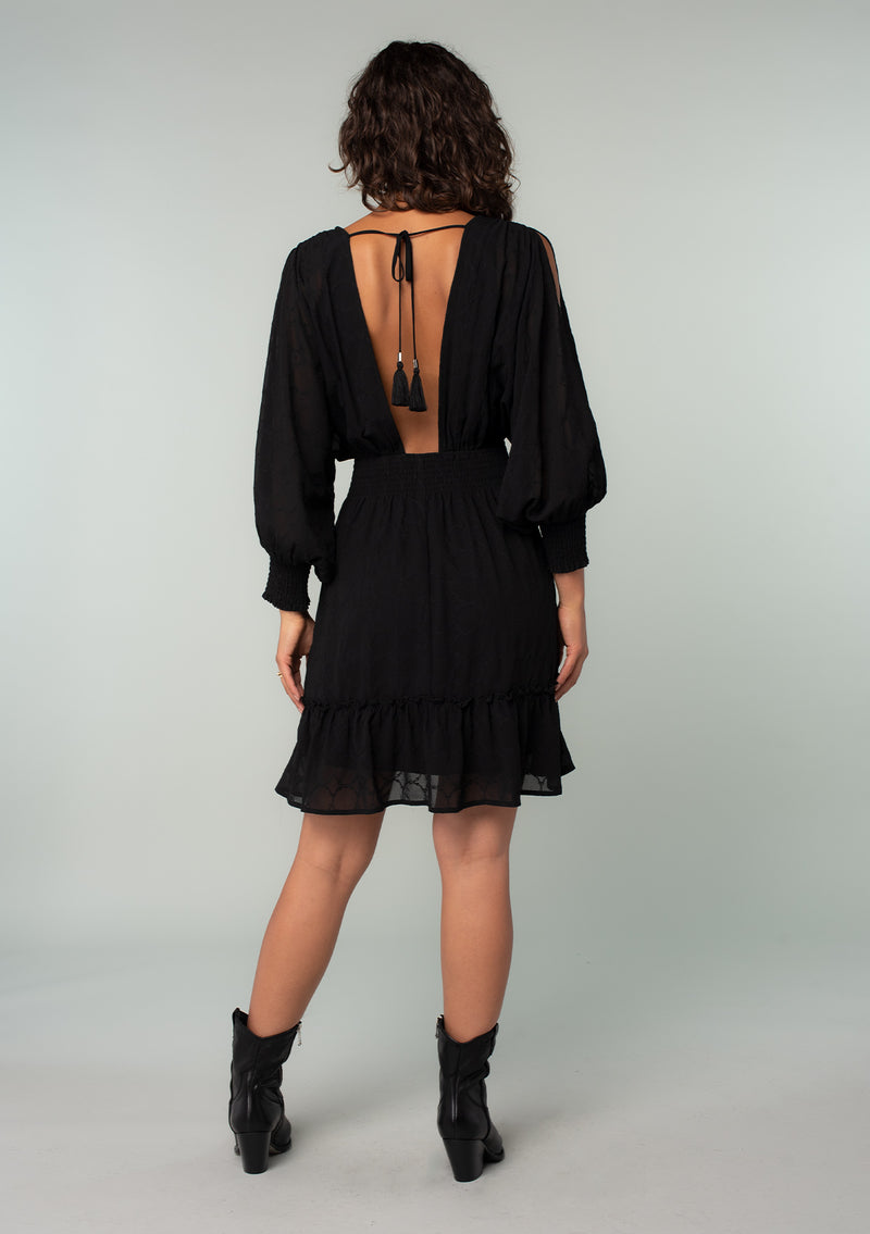 [Color: Black] A back facing image of a brunette model wearing a black bohemian mini dress in embroidered chiffon. With long split sleeves, a ruffle trimmed tiered skirt, a smocked elastic waist, a v neckline, and an open back with tassel tie closure. Perfect for weddings or date nights.
