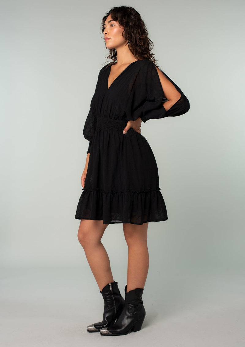 [Color: Black] A side facing image of a brunette model wearing a black bohemian mini dress in embroidered chiffon. With long split sleeves, a ruffle trimmed tiered skirt, a smocked elastic waist, a v neckline, and an open back with tassel tie closure. Perfect for weddings or date nights.