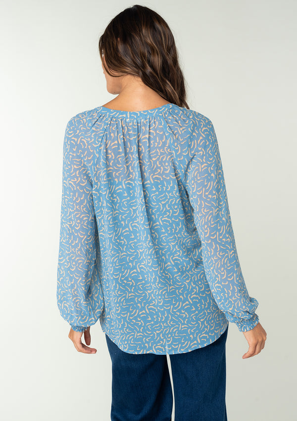 [Color: Dusty Blue/Natural] A back facing image of a brunette model wearing a sheer chiffon blouse in a blue and natural squiggle print. With long sleeves, a self covered button front, a round neckline with split v detail, and smocked elastic wrist cuffs. 