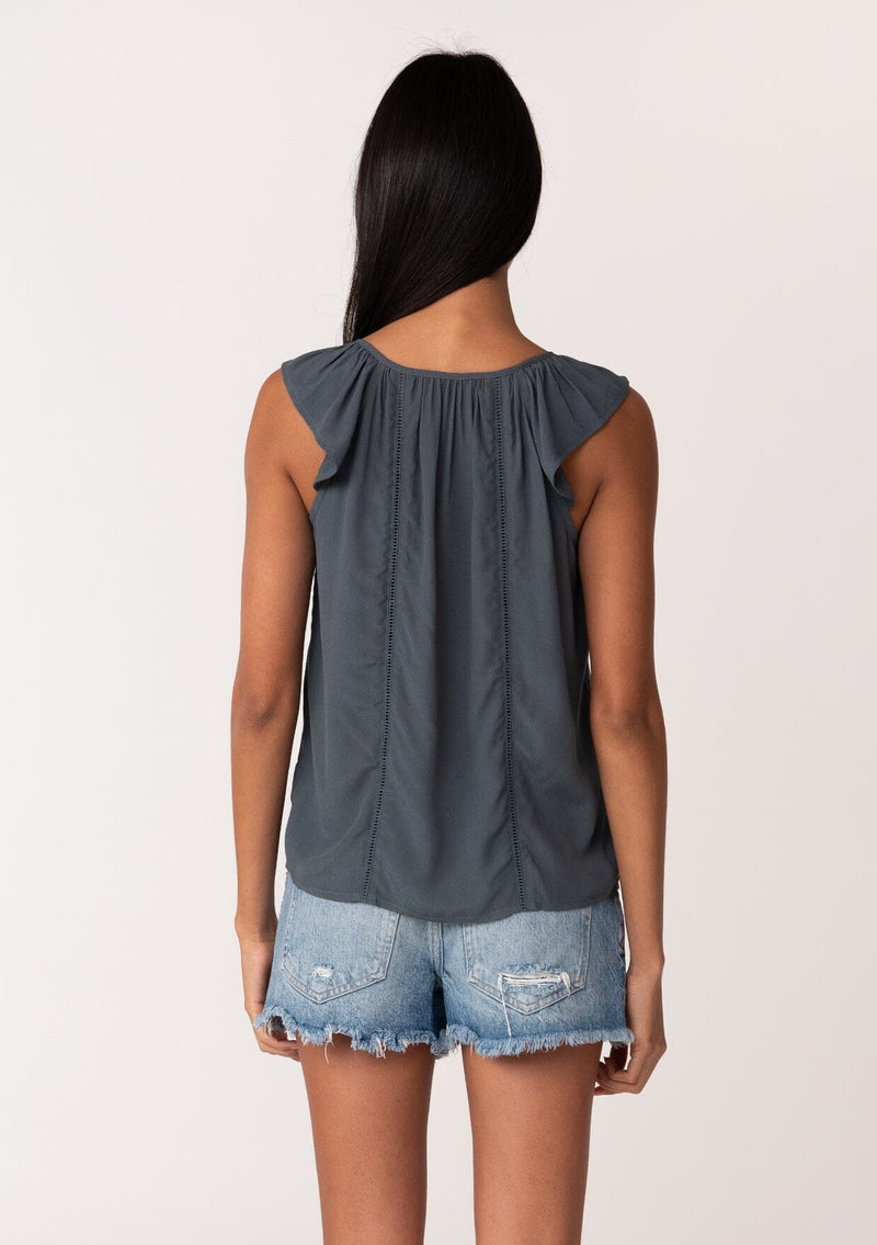 [Color: Slate] A back facing image of a brunette model wearing a slate blue summer top with short flutter straps, a scoop neckline, a button front, and lattice trim. 