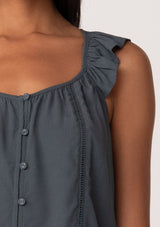 [Color: Slate] A close up front facing image of a brunette model wearing a slate blue summer top with short flutter straps, a scoop neckline, a button front, and lattice trim. 