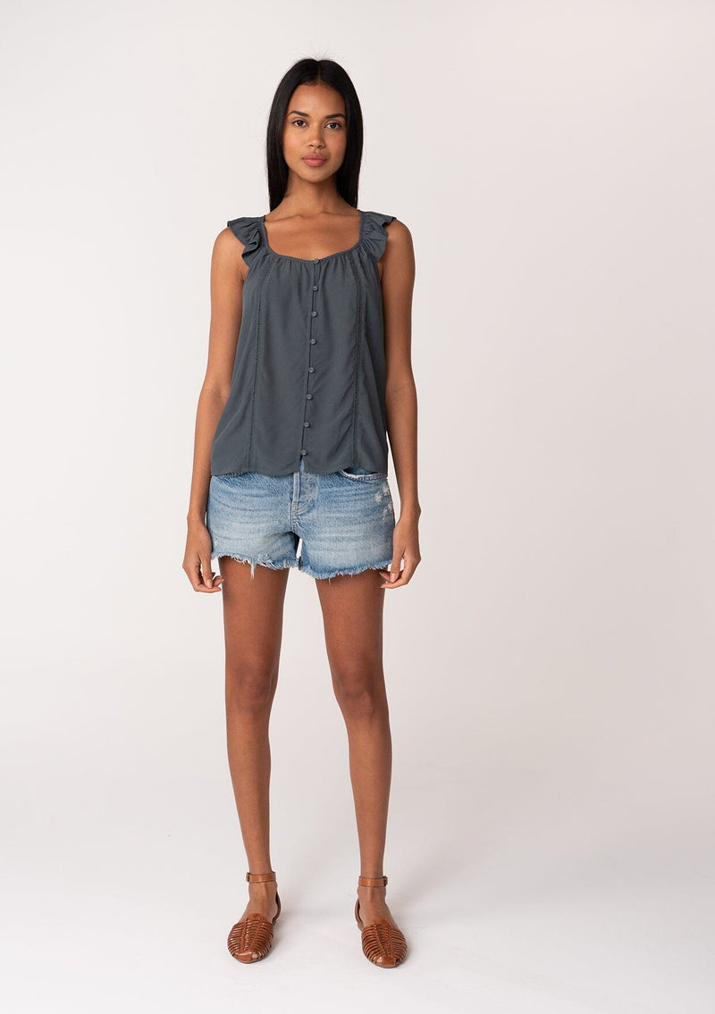 [Color: Slate] A full body front facing image of a brunette model wearing a slate blue summer top with short flutter straps, a scoop neckline, a button front, and lattice trim. 