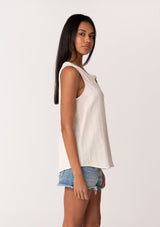 [Color: Cloud] A side facing image of a brunette model wearing a simple cotton off white tank top with a button front, a v neckline, and a relaxed fit. 
