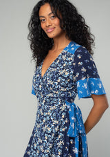 [Color: Navy/Coral] A close up side facing image of a brunette model wearing a navy blue and coral mixed floral print maxi dress. With a wrap silhouette, a side tie closure, a short flutter sleeve, and a v neckline. 