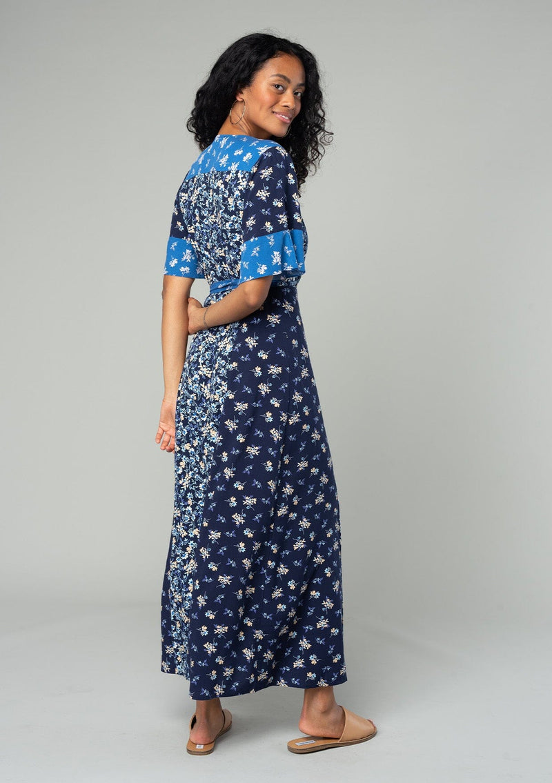 [Color: Navy/Coral] A back facing image of a brunette model wearing a navy blue and coral mixed floral print maxi dress. With a wrap silhouette, a side tie closure, a short flutter sleeve, and a v neckline. 