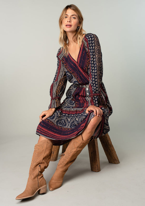 [Color: Navy/Red] A sitting front facing image of a blonde model wearing a bohemian maxi dress in a navy blue and red mixed floral border print. With voluminous flared long sleeves, adjustable wrist ties, a surplice v neckline with hook and eye closure, and an elastic waist. 