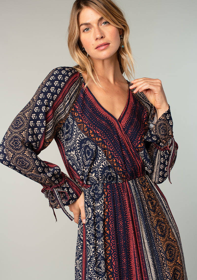 [Color: Navy/Red] A close up front facing image of a blonde model wearing a bohemian maxi dress in a navy blue and red mixed floral border print. With voluminous flared long sleeves, adjustable wrist ties, a surplice v neckline with hook and eye closure, and an elastic waist. 