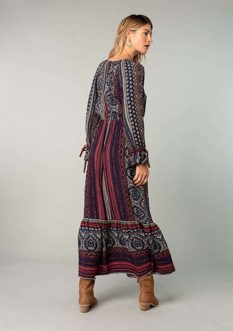 [Color: Navy/Red] A back facing image of a blonde model wearing a bohemian maxi dress in a navy blue and red mixed floral border print. With voluminous flared long sleeves, adjustable wrist ties, a surplice v neckline with hook and eye closure, and an elastic waist. 