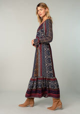 [Color: Navy/Red] A side facing image of a blonde model wearing a bohemian maxi dress in a navy blue and red mixed floral border print. With voluminous flared long sleeves, adjustable wrist ties, a surplice v neckline with hook and eye closure, and an elastic waist. 