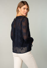 [Color: Navy] A back facing image of a blonde model wearing a navy blue holiday blouse in embroidered chiffon. With three quarter length sleeves, a flutter wrist cuff, self covered button front, and a flowy fit.