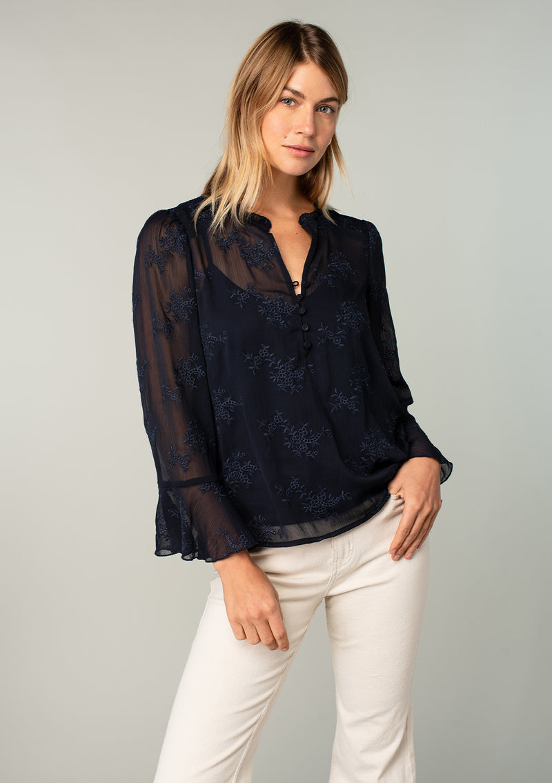 [Color: Navy] A half body front facing image of a blonde model wearing a navy blue holiday blouse in embroidered chiffon. With three quarter length sleeves, a flutter wrist cuff, self covered button front, and a flowy fit.