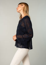 [Color: Navy] A side facing image of a blonde model wearing a navy blue holiday blouse in embroidered chiffon. With three quarter length sleeves, a flutter wrist cuff, self covered button front, and a flowy fit.