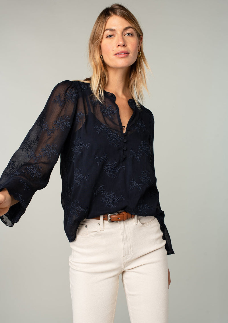 [Color: Navy] A front facing image of a blonde model wearing a navy blue holiday blouse in embroidered chiffon. With three quarter length sleeves, a flutter wrist cuff, self covered button front, and a flowy fit.