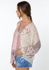 [Color: Natural/Wine] A model wearing a sheer chiffon peasant top in a pink and natural mixed bohemian print. With long voluminous sleeves, a split neckline with tassel ties, and a button front.