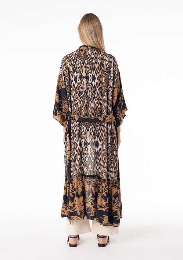 [Color: Black/Taupe] A back facing image of a blonde model wearing a mid length duster lounge robe in a black and taupe mixed floral and ikat print. With half length long sleeves, a ruffle trimmed tiered hemline, an open front, and a tie waist belt.