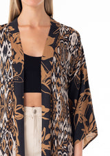 [Color: Black/Taupe] A close up front facing image of a blonde model wearing a mid length duster lounge robe in a black and taupe mixed floral and ikat print. With half length long sleeves, a ruffle trimmed tiered hemline, an open front, and a tie waist belt. 
