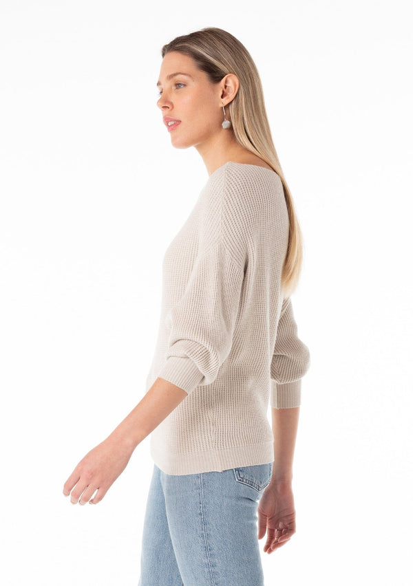 [Color: Heather Stone] A side facing image of a blonde model wearing an off white, stone grey waffle knit pullover sweater. With long sleeves, a relaxed fit, and a wide neckline that can be worn off the shoulder. 