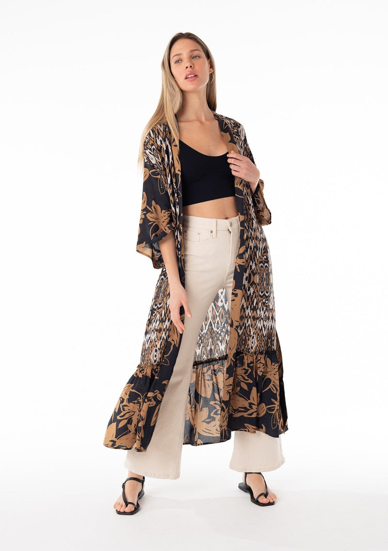 [Color: Black/Taupe] A front facing image of a blonde model wearing a mid length duster lounge robe in a black and taupe mixed floral and ikat print. With half length long sleeves, a ruffle trimmed tiered hemline, an open front, and a tie waist belt. 