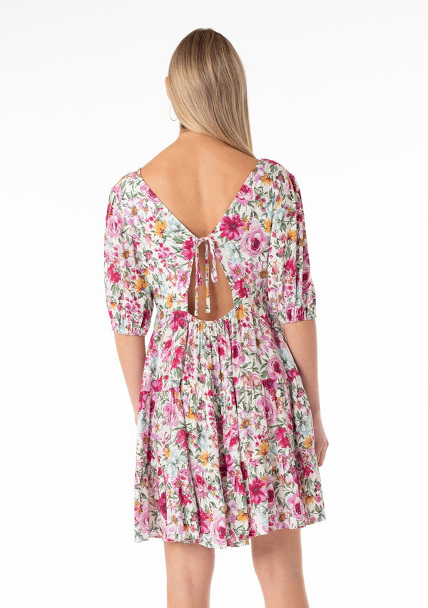 [Color: Natural/Plum] A back facing image of a blonde model wearing a flowy spring mini dress in a pink floral print. With short puff sleeves, a tiered skirt, an empire waist, a v neckline in front and back, and an open back with tie closure. 