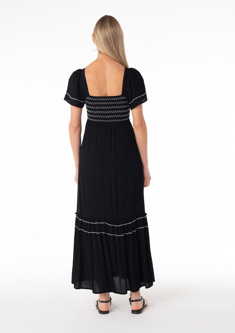 [Color: Black/Ivory] A back facing image of a blonde model wearing a black bohemian maxi dress. With short sleeves, a slim fit smocked bodice, a square neckline in the front and back, a ruffle trimmed tiered skirt, side pockets, and contrast embroidered stitched detail. 