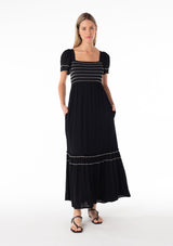 [Color: Black/Ivory] A front facing image of a blonde model wearing a black bohemian maxi dress. With short sleeves, a slim fit smocked bodice, a square neckline in the front and back, a ruffle trimmed tiered skirt, side pockets, and contrast embroidered stitched detail. 