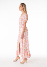 [Color: Peach/Light Pink] A side facing image of a blonde model wearing a classic spring bohemian maxi dress in a peach pink floral print. With short puff sleeves, a front slit, an elastic waist, and a drawstring v neckline with adjustable tie. 