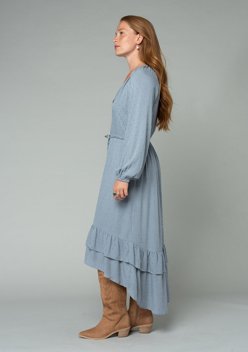 [Color: Grey] A side facing image of a red headed model wearing a light blue grey high low maxi dress with a ruffled hemline, voluminous long sleeves, and a drawstring tassel tie waist.