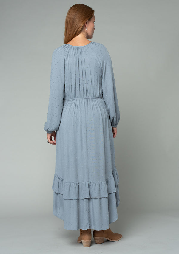 [Color: Grey] A back facing image of a red headed model wearing a light blue grey high low maxi dress with a ruffled hemline, voluminous long sleeves, and a drawstring tassel tie waist.
