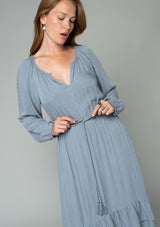[Color: Grey] A close up front facing image of a red headed model wearing a light blue grey high low maxi dress with a ruffled hemline, voluminous long sleeves, and a drawstring tassel tie waist.