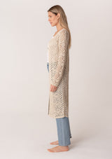 [Color: Natural] A side facing image of a blonde model wearing a classic bohemian cream colored mid length crochet cardigan.