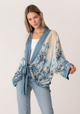 [Color: Blue/Natural] A half body front facing image of a blonde model wearing a bohemian kimono top in a blue floral print. With long flared sleeves, a relaxed flowy fit, and a tie front waist. 