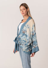 [Color: Blue/Natural] A side facing image of a blonde model wearing a bohemian kimono top in a blue floral print. With long flared sleeves, a relaxed flowy fit, and a tie front waist. 