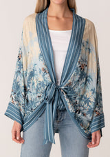 [Color: Blue/Natural] A close up front facing image of a blonde model wearing a bohemian kimono top in a blue floral print. With long flared sleeves, a relaxed flowy fit, and a tie front waist. 