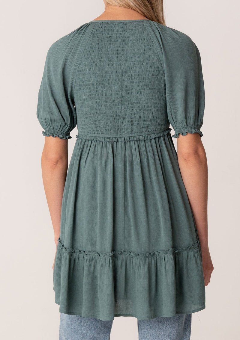 [Color: Lagoon] A back facing image of a blonde model wearing a bohemian spring tunic top in a teal crinkled fabric. With short puff sleeves, ruffle trim, a v neckline, a smocked bodice, an empire waist, and a flowy tiered body. 
