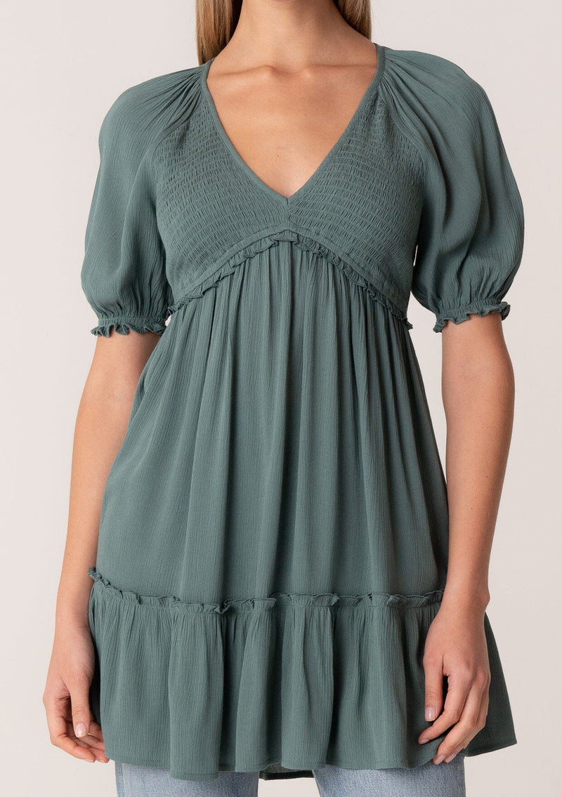 [Color: Lagoon] A close up front facing image of a blonde model wearing a bohemian spring tunic top in a teal crinkled fabric. With short puff sleeves, ruffle trim, a v neckline, a smocked bodice, an empire waist, and a flowy tiered body. 